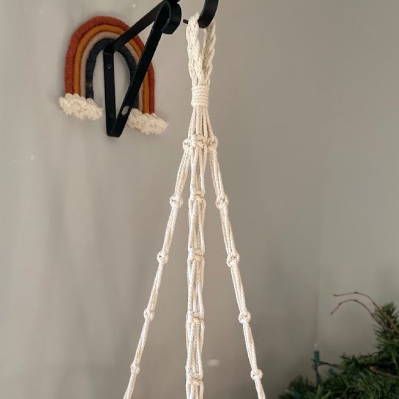 May include: A cream-colored macrame plant hanger suspended from a black metal hook. The plant hanger is knotted with a series of decorative knots. A felt rainbow in shades of orange, yellow, and blue is mounted on the wall.