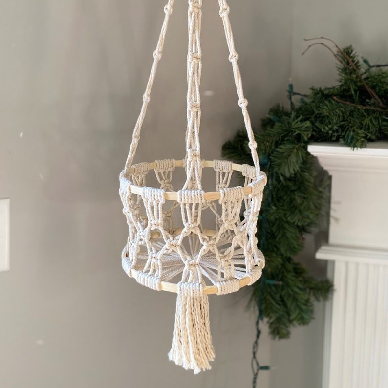 May include: Cream-colored macrame plant hanger with a woven basket design. The plant holder features a wooden frame and a long tassel at the bottom. The hanging plant holder is suspended from a knotted rope.