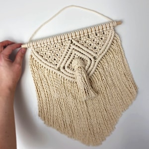May include: A beige macrame wall hanging with a wooden dowel at the top and a tassel at the bottom. The wall hanging is in the shape of a triangle with a geometric pattern.