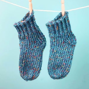 May include: A pair of teal blue knit socks featuring a ribbed cuff and a speckled texture with small flecks of red, yellow, and orange yarn throughout. The socks are hanging from a white string by two wooden clothespins against a light blue background.