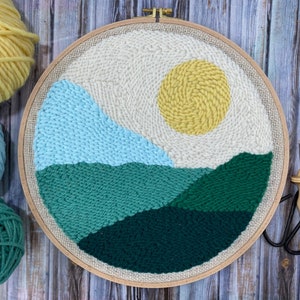 Könnte beinhalten: Ein runder Nadelfilz-Reifen mit einer Landschaftsszene. Die Szene zeigt eine gelbe Sonne, blaue Berge und grüne Hügel. Der Reifen ist aus Holz und hat eine natürliche Oberfläche.