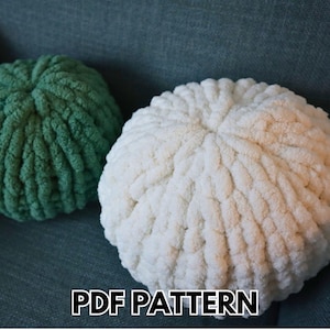 May include: Two round, textured pillows. One is a soft green, the other is white. The pillows are made of a thick, chunky yarn. The text "PDF PATTERN" is displayed in white letters at the bottom of the image.