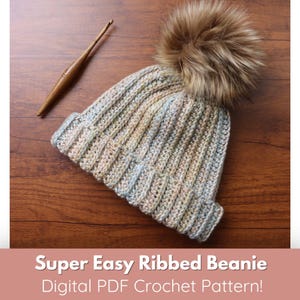 May include: A crochet beanie with a brown faux fur pom pom. The beanie is a light blue, pink, and beige colour. The beanie is on a wooden surface with a crochet hook.
