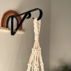 May include: A black metal plant hanger with a cream-colored macrame plant holder. A colorful rainbow wall hanging is mounted on the wall behind the plant hanger. The rainbow features shades of brown, orange, and cream.
