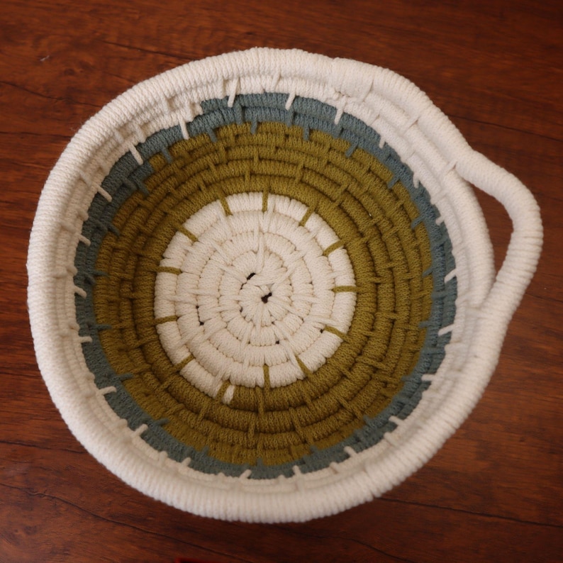 May include: A round, woven basket with a white exterior, a gray-green middle band, and an olive-green inner band. The basket has a white handle and is made of a thick, textured material. The basket is sitting on a wooden surface.