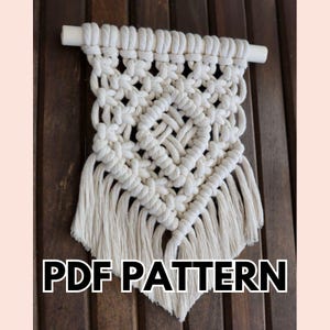 May include: Cream-coloured macrame wall hanging with a geometric design and fringe. The macrame is attached to a white wooden dowel. The text "PDF PATTERN" is displayed in a bold, white font at the bottom of the image.