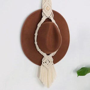 May include: A white macrame hat hanger with a brown hat hanging from it. The hanger has a tassel at the bottom and a wooden bead at the top.