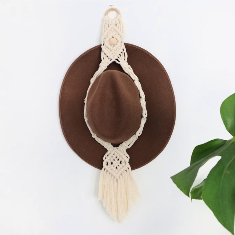 May include: A white macrame hat hanger with a wooden bead and a brown fedora hat hanging from it.