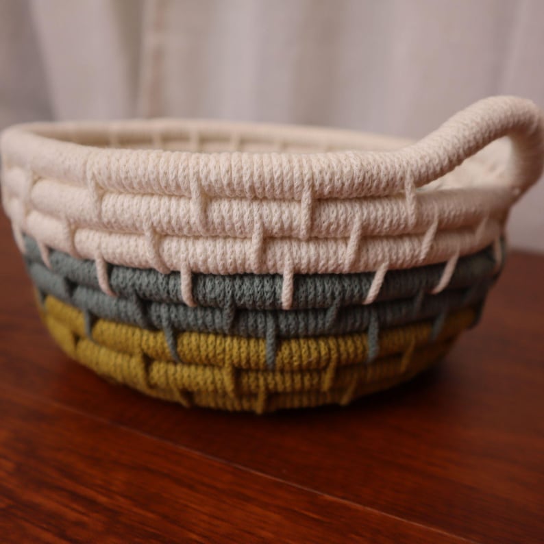May include: A round, woven basket with a handle. The basket features a cream-colored top, a gray-green middle section, and a yellow-green base. The basket is made of a thick, textured material and sits on a wooden surface.