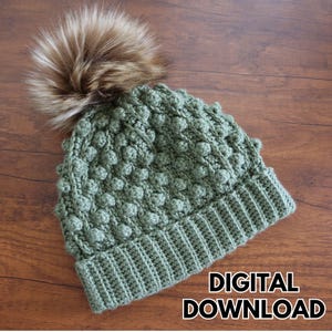 May include: A sage green knitted hat with a textured bobble stitch design and a faux fur pom-pom. The hat has a ribbed brim and the words "DIGITAL DOWNLOAD" in a white font.