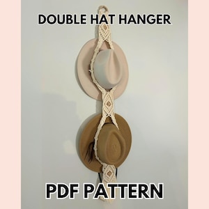 May include: A macrame double hat hanger with two hats. The top hat is light beige, and the bottom hat is brown. The hanger is made of cream-colored woven rope. Text on the image reads "DOUBLE HAT HANGER" and "PDF PATTERN."