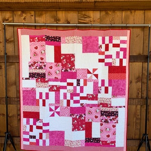 May include: A pink and red patchwork quilt with a variety of heart patterns. The quilt is hanging on a wooden stand.