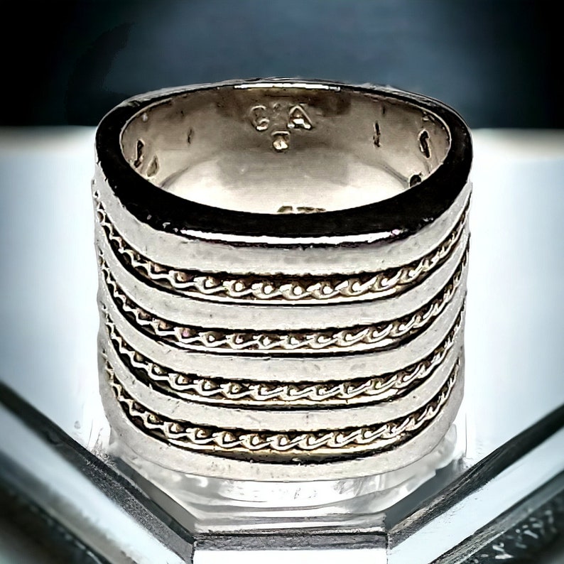May include: A silver ring with a wide band and a textured design. The band is made up of multiple rows of small, linked chains. The ring is marked with the letters "CA" on the inside.