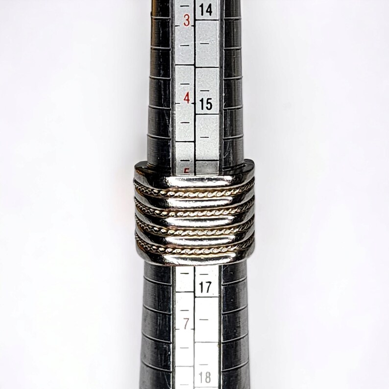 May include: A silver and gold ring with a braided design, measured on a ring sizer with markings from size 14 to 18.