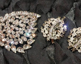 May include: Two silver brooches with clear rhinestone embellishments. The larger brooch is a cluster of rhinestones in a floral shape. The smaller brooch is a cluster of rhinestones in a geometric shape.