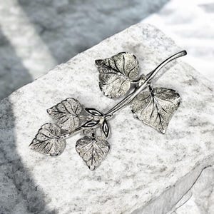 May include: Silver tone brooch featuring a branch design with five textured leaves. The leaves have a slightly raised design.