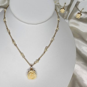 May include: A delicate gold necklace with a white rose pendant and small pearl accents. The necklace is displayed on a white stand.