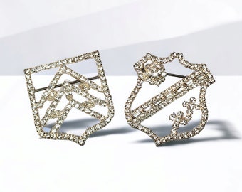 May include: Two silver-toned brooches with a geometric design. Both brooches are covered in clear rhinestones.