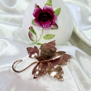 May include: A rose gold-toned metal leaf-shaped hair clip with a hook on the side. The clip is on a white satin background.