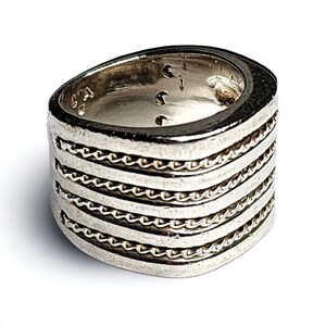 May include: A silver ring with five bands of braided detail. The ring is wide and has a rounded shape.