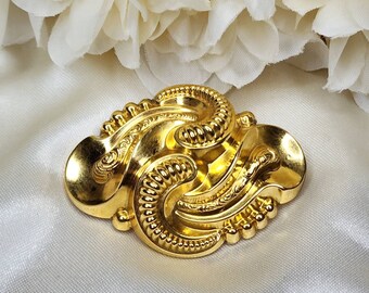 May include: A gold-toned brooch with an intricate design of swirling lines and textured details.
