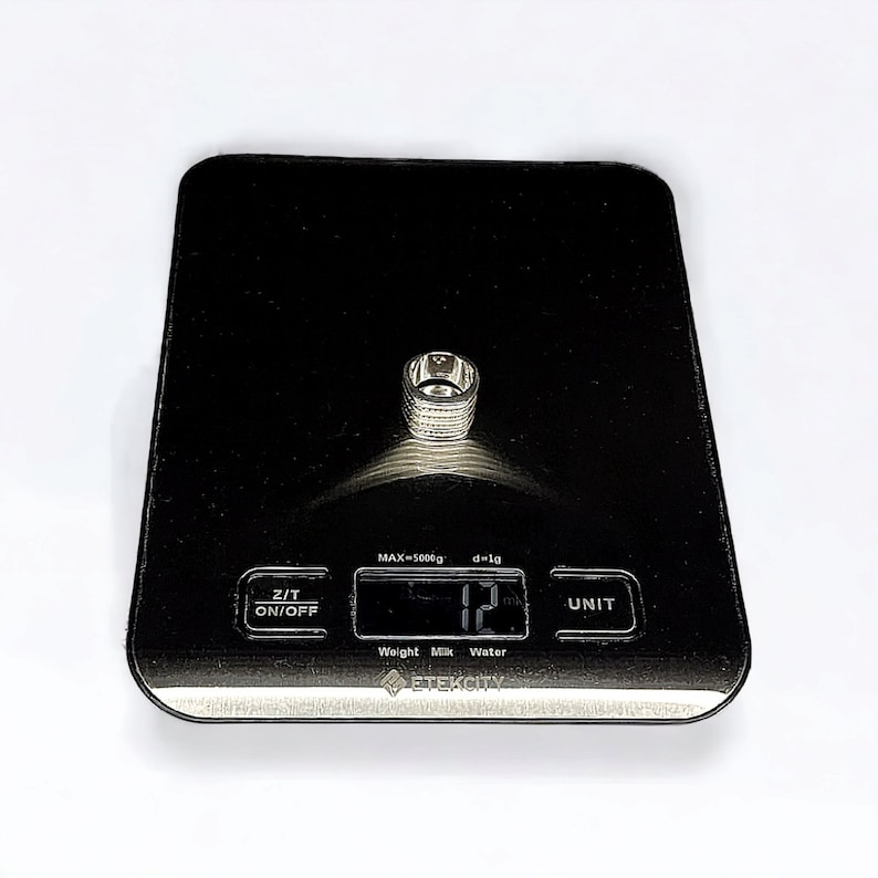 May include: A silver ring on a black digital scale that reads 12 grams. The scale has a black background with white text that reads "MAX=5000g d=1g", "Z/T ON/OFF", "Weight Milk Water", "ETEKCITY", and "UNIT".