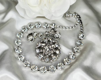 May include: A sparkling rhinestone necklace, brooch, and earrings set. The necklace and earrings feature large, round rhinestones, while the brooch has a more intricate design with smaller rhinestones.