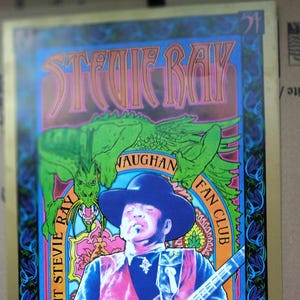 May include: A colorful poster featuring a portrait of Stevie Ray Vaughan, a guitarist, with a bright blue background and a green dragon in the top left corner. The poster has the text "Stevie Ray Vaughan Fan Club" and "The Detroit Stevie Ray Vaughan" in a decorative font.