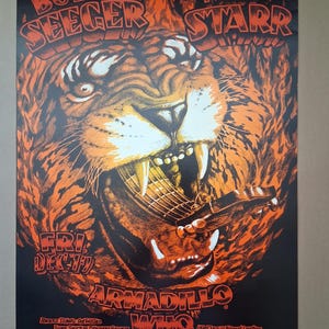 May include: A vintage concert poster featuring a tiger's head with an open mouth, holding a guitar. The poster has orange and black colors with the names "BOB SEEGER" and "RUBY STARR" at the top. The text "FRI DEC 17" and "ARMADILLO WHO" are also visible.