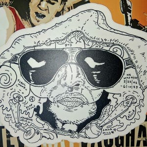 May include: A black and white sticker featuring a stylized illustration of a man wearing sunglasses. The sticker includes the text "HE WAS RIGHT 5/22/42 - 6/10/23" and other text. The design has a cartoonish style.