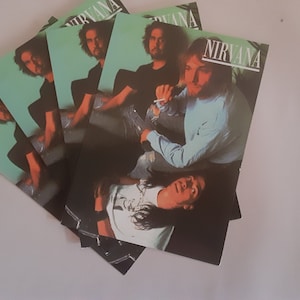 May include: A stack of four Nirvana album covers with a green background and a band photo. The band members are wearing casual clothing and the album title "Nirvana" is printed in white on the top right corner of the cover.