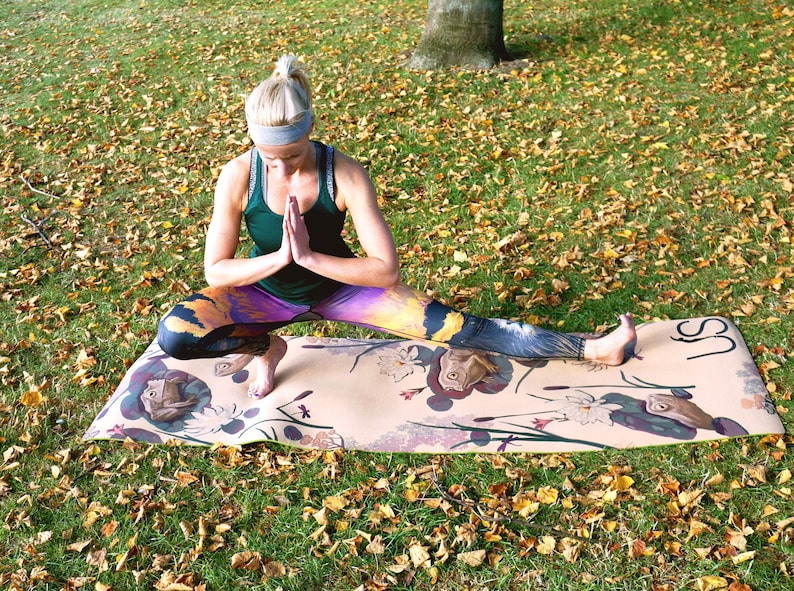 May include: A woman in a green tank top and colorful leggings is doing yoga on a patterned yoga mat outdoors. The mat has a light brown background with a floral design and the word "US" printed on the right side.