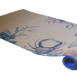 May include: A rolled-up yoga mat with a blue border and a light brown surface. The mat features a watercolour design of blue and pink sea life, including a crab, coral, and seaweed.