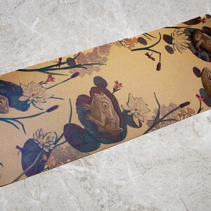 May include: A brown yoga mat with a repeating pattern of frogs, lily pads, and flowers. The mat has the word "US" printed on the bottom left corner.