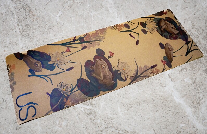 May include: A brown yoga mat with a repeating pattern of frogs, lily pads, and flowers. The mat has the word "US" printed on the bottom left corner.