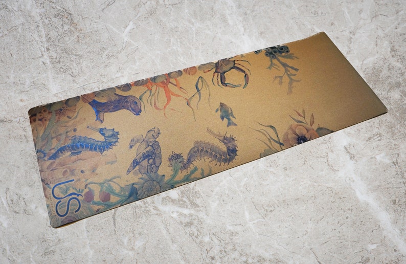 May include: A brown yoga mat with a sea life design. The mat features seahorses, a crab, a turtle, and seaweed. The text "US" is printed on the bottom left corner of the mat.