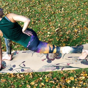 May include: A woman in a green tank top and colorful leggings is stretching on a patterned yoga mat. The mat has a floral design and the text "US" printed on it.