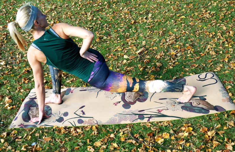 May include: A woman in a green tank top and colorful leggings is stretching on a patterned yoga mat. The mat has a floral design and the text "US" printed on it.