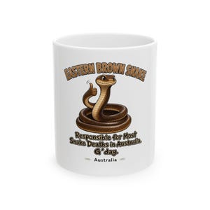 Puede incluir: Una taza de cerámica blanca con una ilustración de dibujos animados de una serpiente marrón enroscada en círculo, haciendo un gesto de pulgar hacia arriba. El texto dice: EASTERN BROWN SNAKE, Responsible for Most Snake Deaths in Australia. G'day. Australia.