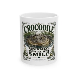 Könnte beinhalten: Ein weißer Keramikbecher mit einer detaillierten Illustration eines Krokodils, das aus dem Wasser auftaucht. Der Text lautet CROCODILE AUSTRALIA'S MOST PATIENT SMILE. Das Design umfasst grüne Schilfhalme, dekorative Ränder und eine kleine Australienkarte.