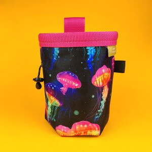 Könnte beinhalten: Ein schwarzer Stoff-Chalkbag mit einem lebendigen Quallenmuster in Pink, Orange, Blau und Grün. Die Tasche hat einen pinkfarbenen Rand, eine pinkfarbene Schlaufe und eine schwarze Schlaufe. Ein schwarzer Kordelzug mit Knebel ist sichtbar.