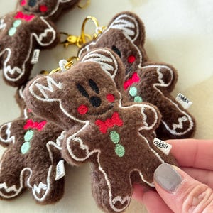 May include: Brown gingerbread man keychains with white icing details, red bowties, and green buttons. The keychains have gold-colored clasps and small white tags with the word "rakkii". A hand is holding one of the keychains.