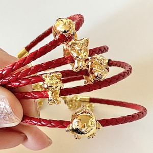 May include: A red braided leather bracelet with gold charms. The charms are shaped like animals, including a pig, a fish, and a bird.