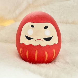 May include: A red and orange traditional Japanese lucky charm, known as a "daruma" doll, with a painted face and a white beard. The doll is sitting on a white fluffy surface.