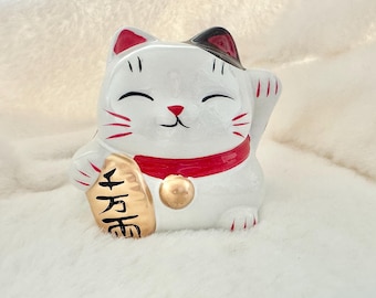 May include: White ceramic Maneki-Neko, or lucky cat, figurine with red and black accents. The cat is holding a gold coin with the Japanese character for "fortune" written on it.