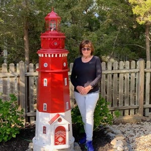 May include: A decorative red and white lighthouse stands tall in a garden setting. The lighthouse features a red lantern room and a white base with a small door. The structure is approximately 6 feet tall and is surrounded by greenery and a wooden fence.