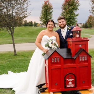 May include: A bride and groom stand in front of a red and black miniature lighthouse. The bride is wearing a white wedding dress and holding a bouquet of white flowers. The groom is wearing a dark suit and a white tie. The lighthouse has a black roof and white trim.