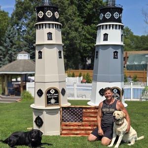 May include: Two decorative lighthouse structures, one beige and one gray, stand on a green lawn. Each has a black top and windows. A wooden American flag and two dogs are in front. The lighthouses have the names "Ken & Janet" and a date.