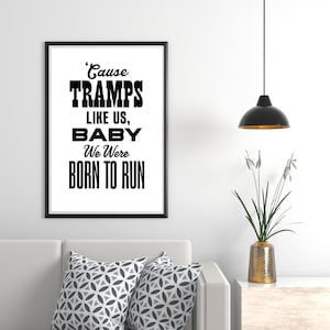 May include: A framed black and white print with the text "'Cause Tramps Like Us, Baby We Were Born To Run." The print hangs above a white sofa with two patterned throw pillows. A black pendant light hangs from the ceiling.