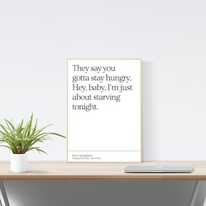 May include: A framed print with a gold border displays the quote "They say you gotta stay hungry. Hey, baby, I'm just about starving tonight." by Bruce Springsteen. The print is on a wooden desk with a plant.